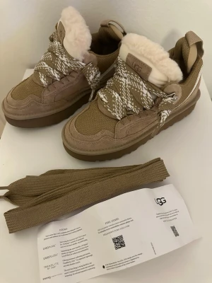 Beige sneakers från UGG med päls - Säljer ett par beige sneakers från UGG med fluffig päls på plösen och insidan. Skorna har grov platt sula, snörning med breda mönstrade skosnören i beige och vitt, samt detaljer i mocka och textil. Perfekt för dig som vill ha både stil och komfort.