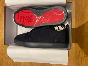 Mörkblå sneakers från Christian Louboutin - Snygga mörkblå sneakers från Christian Louboutin med lyxig röd signatursula och diskret broderad logga på hälen. Skorna har rund tå, platt sula och är tillverkade i mocka med matchande snörning. Perfekt för dig som vill sticka ut med exklusiv stil.