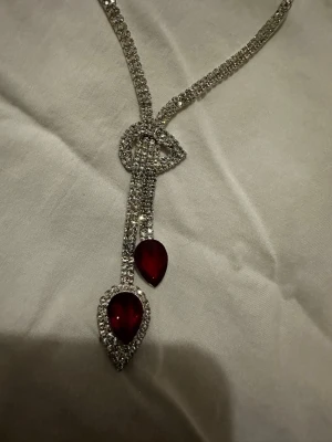 Smyckesset med röda stenar och strass - Säljer ett glittrigt smyckesset med halsband och örhängen. Halsbandet har två droppformade röda stenar omgivna av strass och matchande örhängen. Perfekt för att lyfta din outfit med lite extra bling och färg.