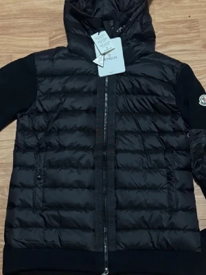 Svart pufferjacka från Moncler - Svart pufferjacka från Moncler med huva och ribbade ärmar. Jackan har quiltad design, dragkedja framtill och Moncler-logga på ärmen. Materialet är polyester och stickad textil på ärmarna. Perfekt för dig som vill ha en snygg och varm jacka med streetkänsla.