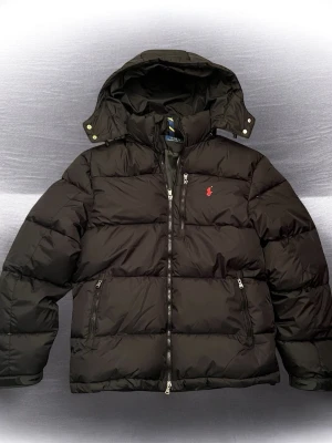 Svart pufferjacka från Polo Ralph Lauren - Svart pufferjacka från Polo Ralph Lauren med röd broderad logga på bröstet. Jackan har huva, flera dragkedjor och två sidofickor med dragkedja. Materialet är polyester och jackan har en quiltad design som ger extra volym och värme.