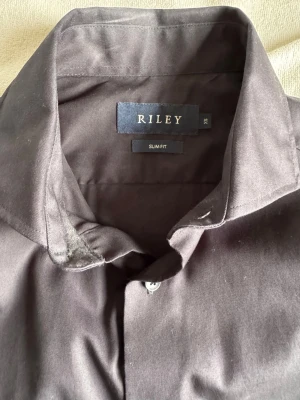 Svart slim fit skjorta från Riley - Svart skjorta från Riley i slim fit-modell, tillverkad i 100% bomull. Klassisk krage och knäppning framtill med svarta knappar. Perfekt för dig som gillar stilrena och enkla plagg med snygg passform.