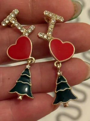 Juliga örhängen med hjärta och gran - Säljer ett par festliga örhängen med gnistrande stenar, röda hjärtan och gröna julgranar. Perfekta accessoarer för att lyfta din look under julen. Örhängena har guldfärgade detaljer och är riktigt söta och unika.