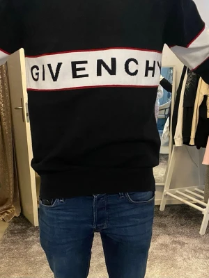Svart stickad tröja från Givenchy - Säljer en svart stickad tröja från Givenchy med vit panel och röd kant över bröstet där loggan är broderad. Tröjan har långärmad passform och en clean, normal siluett. Perfekt för dig som gillar stilrena designer och exklusiva märken. OBS: Storlek XL men sitter som L!