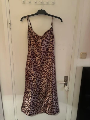 Leopardmönstrad långklänning ( går lite under mina knän och jag  är 161cm lång ) - Säljer en snygg långklänning från LOCH i storlek M med leopardmönster. Klänningen har tunna axelband och en mjuk, glansig yta som ger en lyxig känsla. Färgerna går i beige och bruna toner, och materialet känns lätt och följsamt mot kroppen. Skönt material i sammet. 