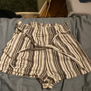 Randiga högmidjade shorts från Divided - Säljer ett par snygga högmidjade shorts från Divided i beige och mörkbrun randigt mönster. De har ett papperspåse-midjeband och matchande bälte i samma tyg. Materialet är 100% bomull, vilket gör dem sköna och luftiga för varma dagar.