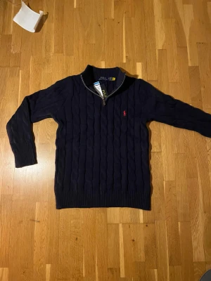 Mörkblå kabelstickad tröja Polo Ralph Lauren - Snygg mörkblå kabelstickad tröja från Polo Ralph Lauren med half zip och klassisk röd broderad logga på bröstet. Tröjan är gjord i 100% bomull och har ribbad krage. Perfekt för dig som gillar stilrena och tidlösa plagg. 