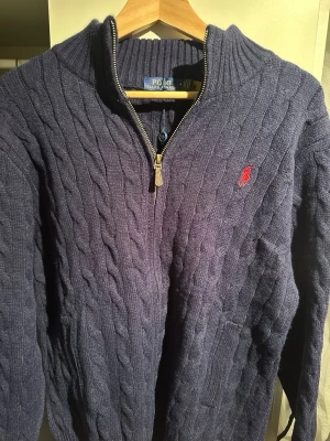 Mörkblå stickad tröja Polo Ralph Lauren - Säljer en mörkblå stickad tröja från Polo Ralph Lauren med half zip och kabelstickat mönster. Tröjan har långärmad design och röd broderad logga på bröstet. Perfekt för dig som gillar klassisk och stilren look.