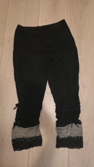 Svarta trekvartsleggings  - Svarta piratbyxor med svartvitrandiga detaljer, spetsdetaljer samt rynkade detaljer på utsida ben. Nakenkatt finns i hemmet