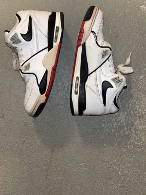 Nike Air Flight vita sneakers - Nike Air Flight sneakers i vitt med svarta och röda detaljer. Skorna har snörning, synlig Air-enhet i sulan och klassisk Nike-logga på sidan och hälen. Ovandelen är i skinn med mesh vid tungan för extra komfort. Snygg retrostil med rund tå och platt sula.