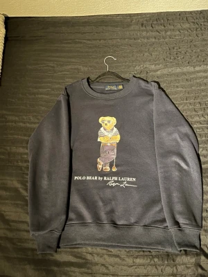 Svart sweatshirt Polo Bear Ralph Lauren - Mörk Blå sweatshirt från Ralph Lauren med Polo Bear-tryck på framsidan. Tröjan har rund hals, ribbade muddar och är långärmad. Materialet är mjuk bomull och polyester, perfekt för chill dagar. Klassisk och stilren design med ikonisk björn. Säljer den då jag bara använt den endast 2 gånger! 