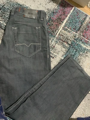 Mörkgrå Boss jeans comfort fit W33 L32 - Snygga mörkgrå jeans från Boss med comfort fit och raka ben. Jeansen har kontrasterande orange sömmar, klassisk femficksdesign och knappgylf. Materialet är slitstarkt jeans och passformen är normal, perfekt för dig som gillar stilrena och bekväma byxor.