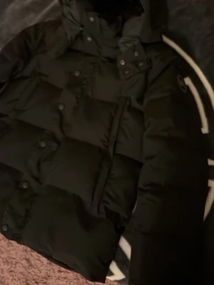 Svart pufferjacka från Canada Goose - Säljer en svart pufferjacka från Canada Goose med avtagbar pälsdetalj till huvan. Jackan har stora quiltade partier, tryckknappar och dragkedja framtill samt en snygg patch på ärmen. Perfekt för kalla dagar och riktigt varm. Passar både s och m i storlek 