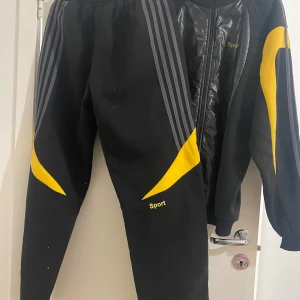 Svart sport jacka  med byxor  - Svart sportväst med blanka paneler framtill och gula inslag på ärmarna. Västen har dragkedja och grå ränder längs axlarna. Perfekt för träning och har en modern, sportig look.