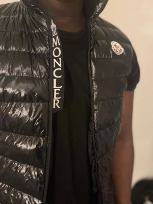 Svart dunväst från Moncler - Säljer en svart, glansig dunväst från Moncler med quiltad design och dragkedja framtill. Västen har ett stort Moncler-tryck på insidan av kragen och klassisk logotyp på bröstet. Insidan har en cool serietidningsetikett och ficka med röd, vit och blå kant.