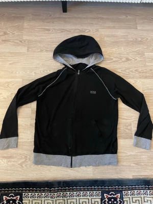 Svart Hugo boss kofta. - Svart hoodie från Hugo Boss med grå detaljer på muddar, nederkant och insida av huvan. Använt rätt mycket.
