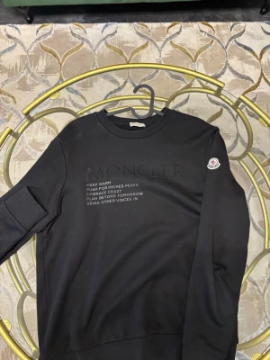 Svart Moncler sweatshirt - Svart sweatshirt från Moncler med tryckt text på bröstet och klassisk Moncler-logga på vänster ärm. Tröjan har rund hals, lång ärm och är gjord i mjuk bomull med insida i fleece. Snygg och enkel design som passar till jeans.