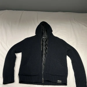Svart stickad hoodiejacka från Replay - Svart stickad hoodiejacka från Replay med quiltat foder och dragkedja framtill. Jackan har två stora fickor, ribbade muddar och en skön huva. Perfekt för dig som gillar en chill och stilren look.