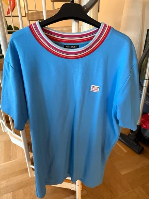 Blå t-shirt med randig krage Acne Studios - Snygg blå t-shirt från Acne Studios med korta ärmar och en fet randig krage i rött, vitt och rosa. Liten broderad patch på bröstet. Materialet är en mix av viskos, nylon och elastan som känns mjukt och skönt mot huden. Perfekt för dig som gillar färg och stil.