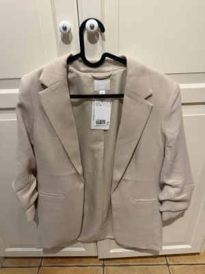 Beige kavaj från H&M - Snygg beige kavaj från H&M med klassisk krage och två fickor framtill och rynkad ärm. Kavajen har en stilren och rak passform med långa ärmar och är tillverkad i ett mjukt material.