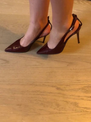 Vinröda klackar med spetsig tå - Helt nya vinröda klackar med spetsig tå och hög klack. Skorna har en öppen häl med rem och är tillverkade i glansigt syntetmaterial. Jättefin vinröd färg.  Köpta utomlands, nypris ca 900