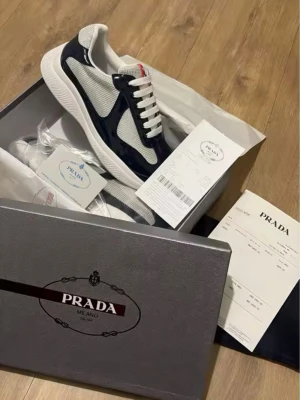 Prada sneakers i vit och svart - Säljer ett par snygga Prada sneakers i vit och marinblå med röda detaljer. Skorna har rund tå, platt sula och snörning. Ovandelen är i mesh och lackad skinn, vilket ger en lyxig och sportig vibe. Kommer med originalkartong, dustbag och oäkthetsbevis.