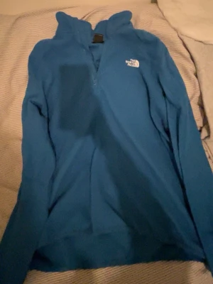 Blå fleece-tröja från The North Face - Säljer en blå fleece-tröja från The North Face med halv dragkedja och hög krage. Perfekt för kyliga dagar och outdoor-aktiviteter. Tröjan har lång ärm och logga broderad på bröstet. Skön och mjuk känsla, passar dig som gillar friluftsliv.