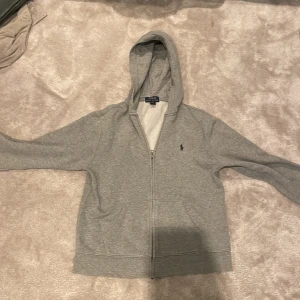 Grå hoodie från Polo Ralph Lauren - Grå hoodie från Polo Ralph Lauren med dragkedja framtill och klassisk broderad logga på bröstet. Tröjan har huva, långa ärmar och är tillverkad i mjuk bomullsmix. Perfekt för chill dagar eller när du vill ha en clean och enkel look.