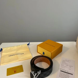 Louis Vuitton svart rutigt skärp - Säljer ett exklusivt Louis Vuitton bälte i svart och grått rutigt mönster med ikoniskt LV-spänne i silver. Bältet är tillverkat i skinn och har detaljer i brunt på insidan. Kommer med originalbox, dustbag och äkthetskort.