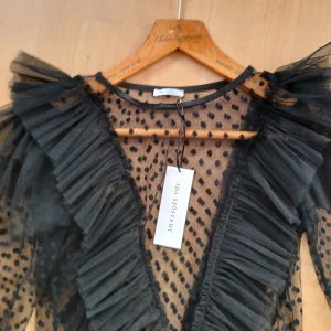 Ny  blus från Ida Sjöstedt - Säljer en transparent svart blus från Ida Sjöstedt i mesh med stora prickar och voluminösa volangdetaljer längs v-ringningen. Blusen har långa puffärmar och är tillverkad i tunn tyll som ger en riktigt statement-look.