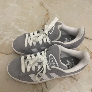 Grå Adidas Campus sneakers - Säljer ett par grå Adidas Campus sneakers med klassiska vita ränder och vit logga på plösen. Skorna har vit sula, rund tå och är i mocka med vita snören. Extra gråa skosnören medföljer. Perfekta för dig som gillar stilren och tidlös streetwear.