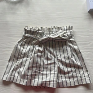Randiga högmidjade shorts från Zara - Snygga högmidjade shorts från Zara i vitt med svarta vertikala ränder. De har papperspåse-midja, knytband i midjan och bälteshällor. Tillverkade i mjuk bomull, perfekta för varma dagar. 
