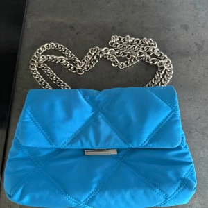 Blå axelväska från Mango - Snygg quiltad axelväska i klarblått från Mango med silverfärgad kedjerem. Väskan har ett mjukt tygmaterial, lockstängning med metallspänne och flera fack inuti. Perfekt för dig som vill ha en färgklick till din outfit.