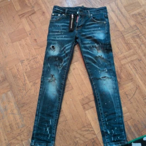 Dsquared2 skinny jeans med dragkedja - Snygga blå skinny jeans från Dsquared2 med slitningar, färgstänk och en stor dekorativ dragkedja framtill. Jeansen har klassisk femficksdesign, låg midja och smal passform. Perfekta för dig som gillar en edgy och trendig look.