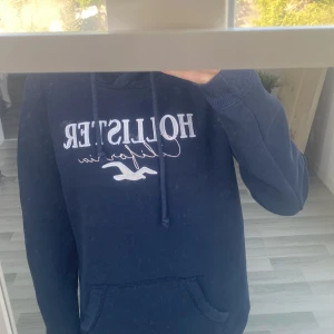 Mörkblå hoodie från Hollister S - Mysig mörkblå hoodie från Hollister med broderad logga och text på bröstet. Tröjan har huva med snörning och en stor magficka. 