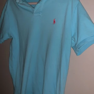Ljusblå Polo Ralph Lauren t-shirt - Snygg ljusblå t-shirt från Polo Ralph Lauren i slim fit-modell. Klassisk pikékrage och korta ärmar, med den ikoniska rosa broderade loggan på bröstet. Tillverkad i mjuk bomull som känns skön mot huden.