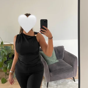 Svart ärmlös topp med gulddetalj - Snygg svart ärmlös topp med en unik guldfärgad detalj på ena axeln. Toppen har en tight passform och är tillverkad i ett mjukt, stretchigt material som sitter skönt mot kroppen. Perfekt för dig som gillar stilrena och trendiga plagg.