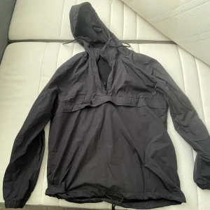 Svart vindjacka från Divided H&M - Svart vindjacka med huva från Divided H&M. Jackan har meshfoder, dragsko i nederkant och halv dragkedja framtill. Lätt och smidig modell med lång ärm, perfekt för blåsiga dagar. Enkel och stilren design.