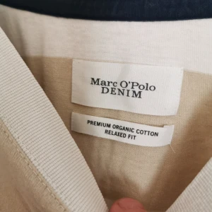 Randig t-shirt från Marc O'Polo Denim - Snygg t-shirt från Marc O'Polo Denim i breda beige och vita ränder. Modellen har korta ärmar, relaxed fit och är gjord av premium ekologisk bomull. Perfekt för dig som gillar en chill och stilren look med lite extra edge.