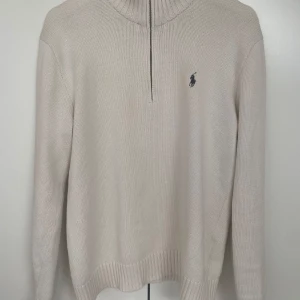 Beige stickad half zip från Polo Ralph Lauren - Snygg beige stickad tröja från Polo Ralph Lauren med half zip och hög krage. Klassisk broderad logga på bröstet och ribbade muddar. Perfekt för lager-på-lager och chill stil.