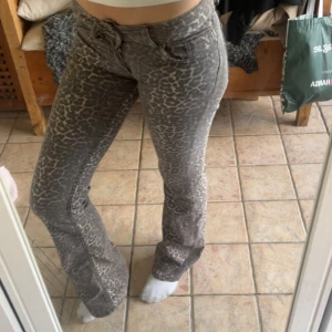 Grå leopardmönstrade bootcut jeans - Säljer ett par grå jeans med leopardmönster och bootcut-ben. Byxorna har låg midja, klassisk femficksdesign och är tillverkade i ett stretchigt jeanstyg. Perfekta för dig som vill sticka ut med ett djurmönster och cool passform.