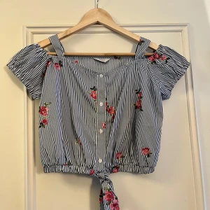 Randig blommig blus från Primark - Söt blus från Primark med smala marinblå och vita ränder, broderade rosa blommor och knytdetalj nedtill. Toppen har korta puffärmar, axelband och knappar framtill. Perfekt för en somrig look!