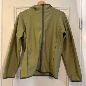 Grön fleecejacka från Quechua - Ljusgrön fleecejacka från Quechua med huva och hel dragkedja framtill. Jackan har två sidofickor med dragkedja och grå detaljer vid kanterna. Perfekt för utomhusaktiviteter och sköna promenader.