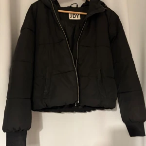 Svart pufferjacka från JDY med huva - Svart pufferjacka från JDY i storlek S. Jackan har en skön huva, dragkedja framtill och ribbade muddar vid ärmsluten. Perfekt för kyliga dagar och har en enkel, stilren look som passar till det mesta.