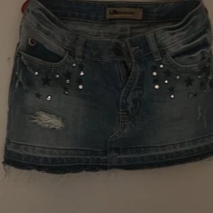 Jeanskjol med stjärnor och nitar - Cool jeanskjol från La Strada med slitna detaljer, stjärn- och nitdekorationer framtill samt fransig nederkant. Klassisk femficksmodell i blå denim, perfekt för dig som gillar en edgy stil.