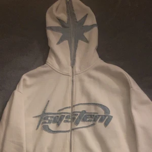 Beige hoodie med blått tryck och stjärna - Säljer en beige hoodie från System med hel dragkedja och stor blå stjärna på huvan. Framsidan har ett broderat blått logotyp-tryck. Hoodien har huva och är perfekt för dig som gillar streetwear vibes.
