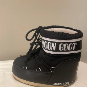 Svarta Moon Boots  - Säljer ett par klassiska svarta Moon Boots med vit logga och snörning framtill. Storlek 37. Använda en vinter och har en liten skada på ena hälen, men inget som syns. Perfekta för vintern och riktigt sköna att ha på sig när det är kallt ute.