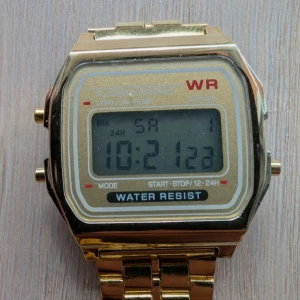 Digital guldfärgad retro-klocka - Säljer en digital retro-klocka i guldfärg med klassisk fyrkantig display och länkarmband. Klockan har digital tidvisning, alarm och stoppursfunktion. Metallarmbandet ger en cool vintagekänsla och displayen är tydlig med svart text mot grå bakgrund.