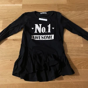 Svart klänning med text No.1 Awesome - Svart långärmad klänning från KappAhl med vit text 'No.1 AWESOME' och små hjärtan framtill. Klänningen har volang nertill och är i mjuk bomull, perfekt för en cool och avslappnad stil.