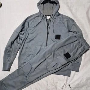 Nike Air Max grå hoodie med dragkedja - Nike Air Max hoodie i grått med svart patch på bröstet, hel dragkedja och huva med snörning. Mjuk och sportig känsla, perfekt för chill eller träning. Långa ärmar och klassisk passform. Snyggt Air Max-tryck på ryggen.